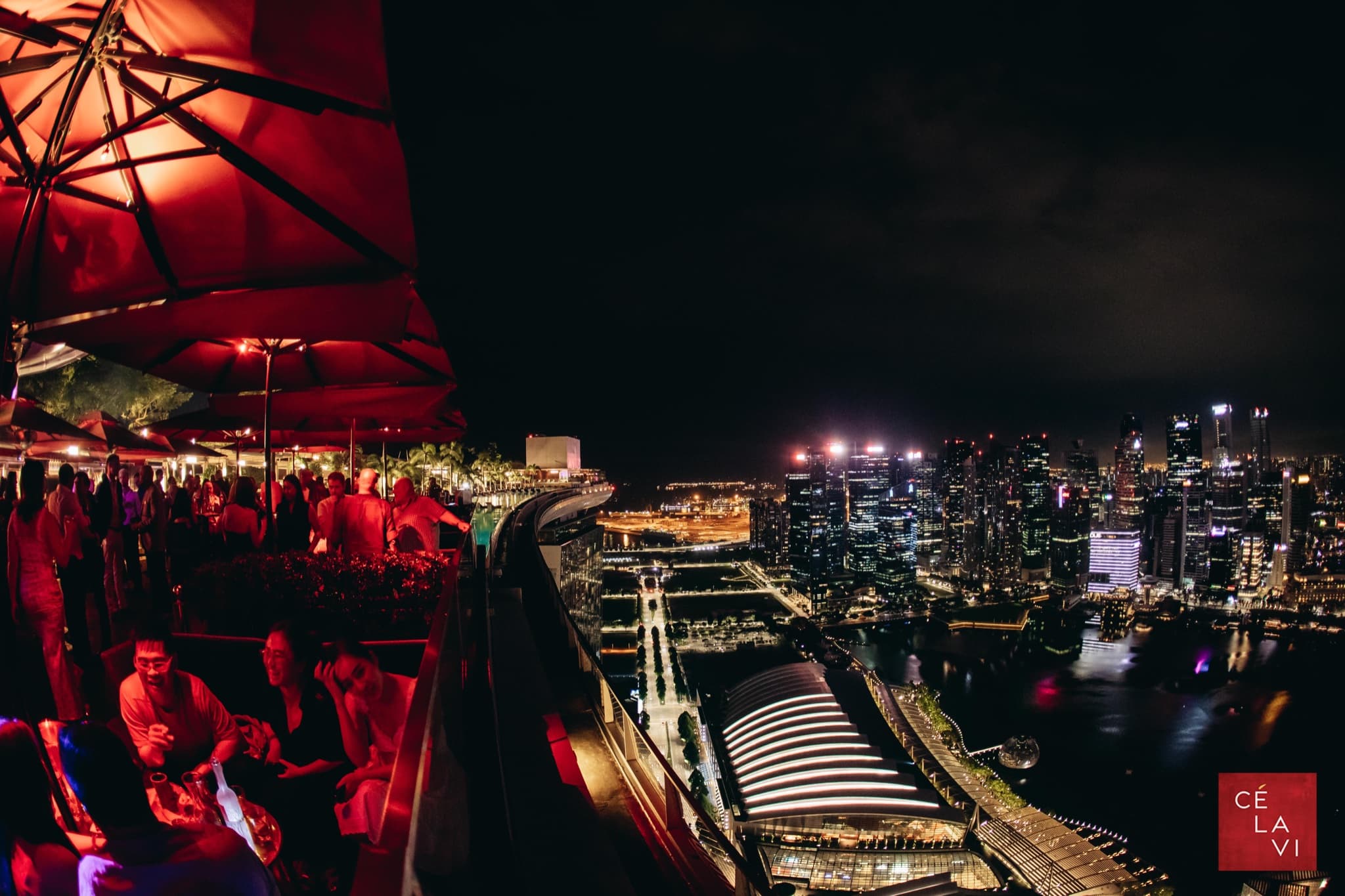 Night view from CÉ LA VI rooftop lounge overlooking Marina Bay during F1 Singapore weekend, with people mingling under red parasols.