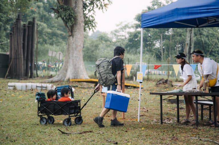 A man carries a icebox and pulls a cart with two children towards the entrance of the festival.
