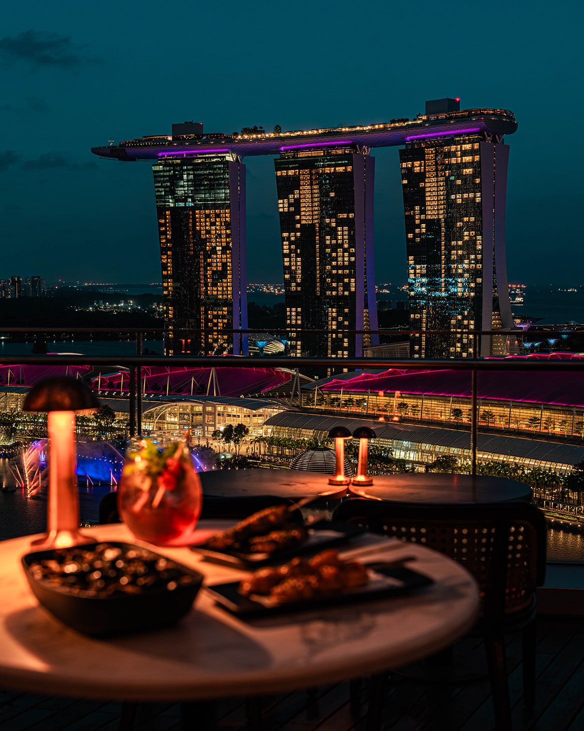 Night view from VUE rooftop bar in Singapore with Marina Bay Sands skyline and happy hour cocktails on the table.