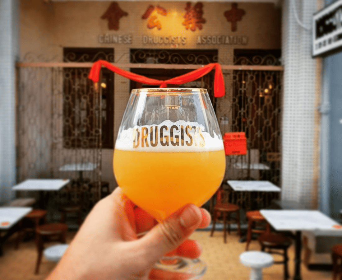 Craft beer in a branded Druggists glass held in front of the historic Chinese Druggists Association, a hidden gem bar in Jalan Besar, Singapore.